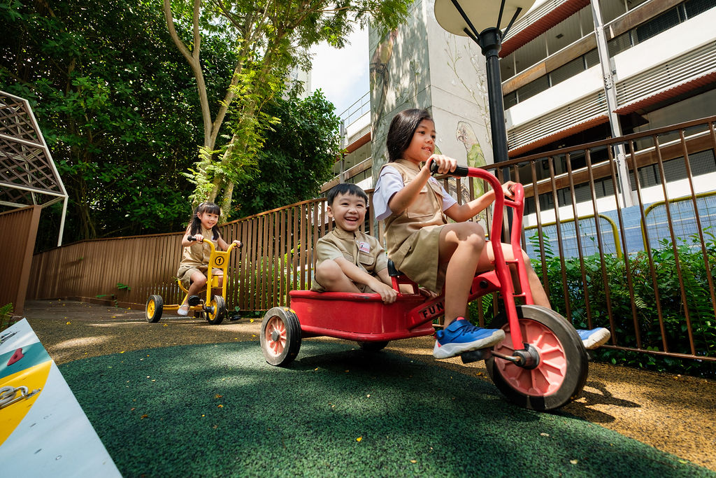 Children riding tricycles down a gentle slope at a playground, smiling and playing together outdoors.